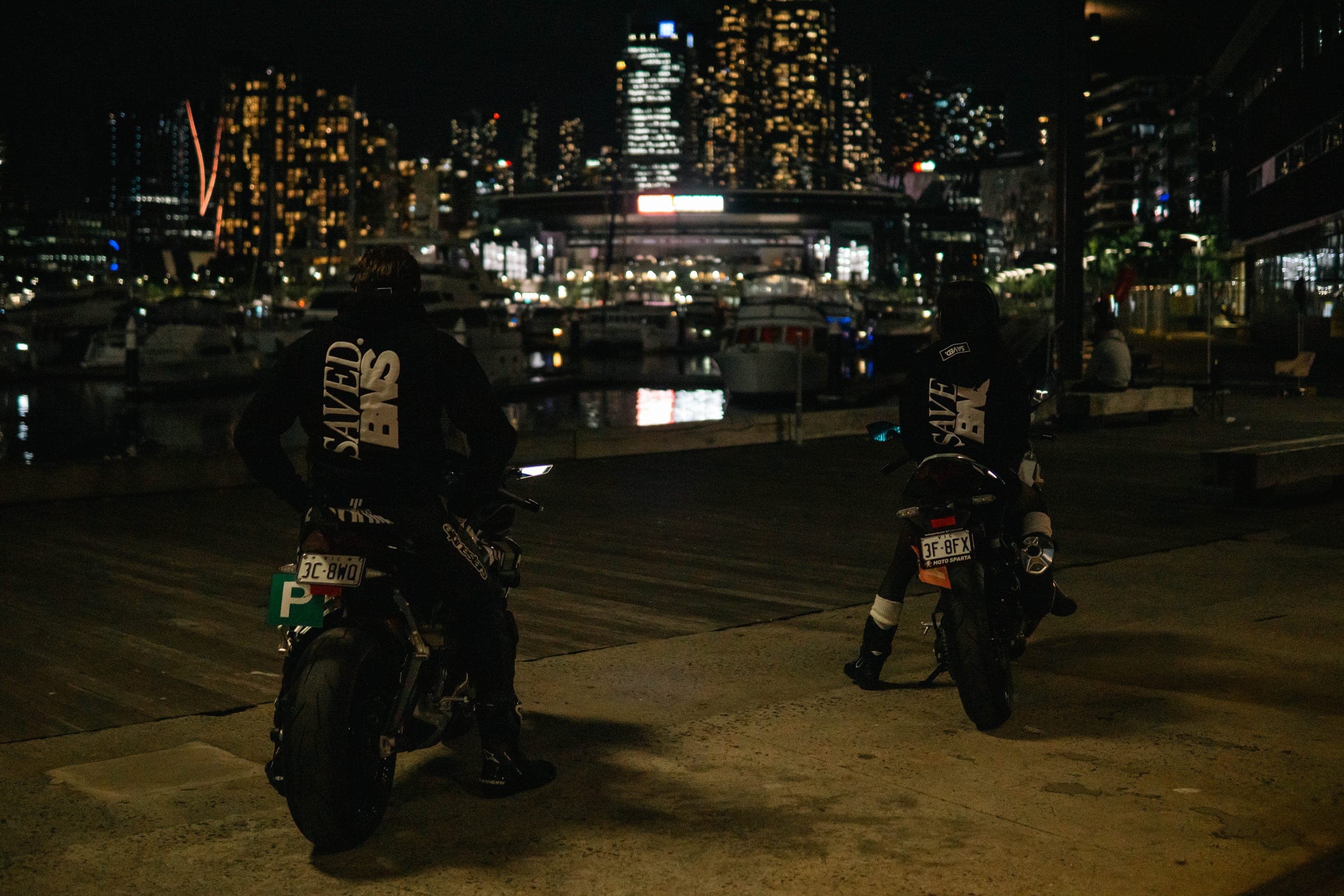 Two riders in SAVED. BNS hoodies stopped on their bikes at Docklands, the Melbourne skyline behind them